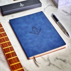 May include: A blue leather-bound journal with a Masonic symbol embossed on the front cover. The symbol features a compass and square with a 'G' in the center. The journal is lying on a white marble surface with a silver pen next to it.