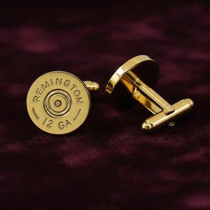 May include: A pair of gold-colored cufflinks. Each cufflink features the text "REMINGTON" and "12 GA" around a central circle. The cufflinks have a shiny finish and a classic design.