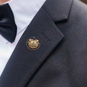 May include: A gold and blue lapel pin with the words "PRINCE HALL" and a Masonic emblem. The pin is round and is affixed to a dark navy blue suit jacket. A black bow tie and white dress shirt are also visible.