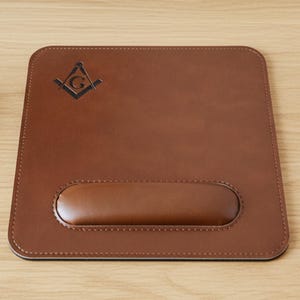 May include: Brown leather mouse mat with an integrated wrist rest. The mat has a stitched border and a Masonic symbol in the upper left corner. The wrist rest is oval and made of the same brown leather.