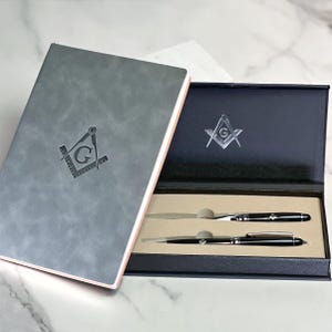 Peut inclure: Un journal relié en cuir gris avec un symbole maçonnique en relief sur la couverture, et une boîte cadeau noire contenant deux stylos argentés avec des symboles maçonniques.