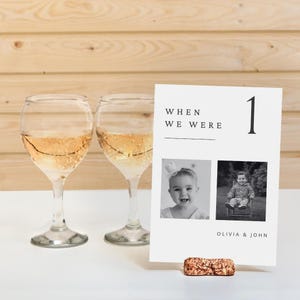 May include: Table number card with a black and white photo of a young girl and boy. The card has the text "When we were" and the number "1" in black text. The card is on a cork stand. The text "Olivia & John" is at the bottom of the card.