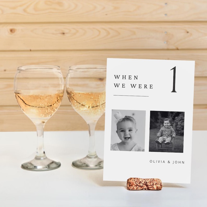 May include: Table number card with a black and white photo of a young girl and boy. The card has the text "When we were" and the number "1" in black text. The card is on a cork stand. The text "Olivia & John" is at the bottom of the card.