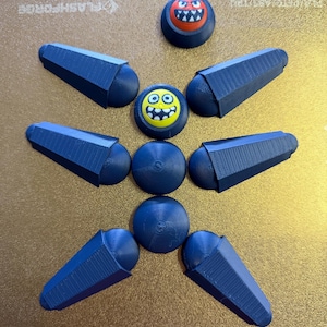 May include: An assortment of 3D-printed items, including blue microphone-shaped objects, round blue pieces, and two circular pieces with cartoon faces. A gray rectangular piece is at the top. The items are arranged on a gold-colored surface.