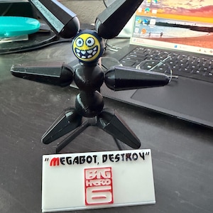 Può includere: Una figura nera di Megabot stampata in 3D da Big Hero 6, con un volto sorridente e arti articolati. La figura è in piedi davanti a una base rettangolare bianca con le parole "MEGABOT, DESTROY" e il logo di Big Hero 6.