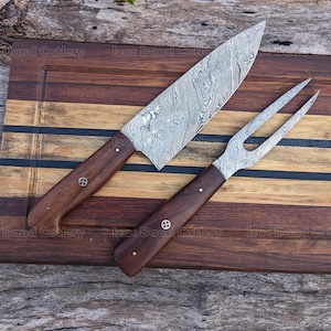 May include: A chef's knife and carving fork set with patterned steel blades and dark wood handles. The knife has a long, pointed blade, and the fork has two tines. The set rests on a wooden cutting board with a striped design. The brand name "Izac Daud Cutlery" is visible.