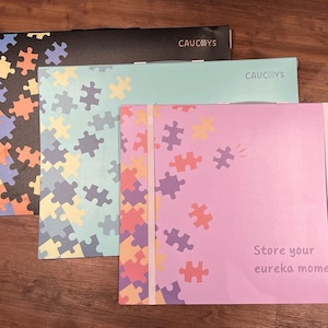 May include: Three pastel-coloured folders with a jigsaw puzzle design and the text "Store your eureka moments" on the pink folder. The folders are labelled "Caucoys" in the top right corner.