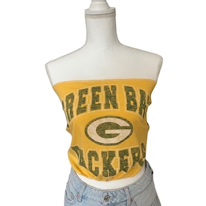 Puede incluir: Un top de tubo amarillo con las palabras "GREEN BAY PACKERS" en letras verdes. El top también presenta el logotipo del equipo. El top se combina con una falda vaquera azul claro. El top se muestra en un maniquí blanco.