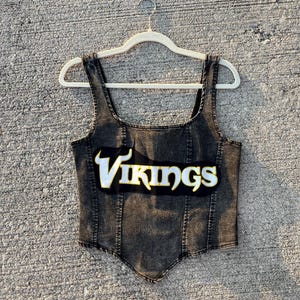 May include: A black denim corset top with the word "Vikings" in white and gold lettering. The top has a square neckline and spaghetti straps. The garment is hanging on a white hanger.
