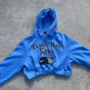 May include: A light blue cropped hoodie featuring the text TAMPA BAY RAYS in black and white lettering. Below the text is a black rectangular patch with a white Nike swoosh logo and the letters TB. The hoodie includes a hood with matching drawstrings.