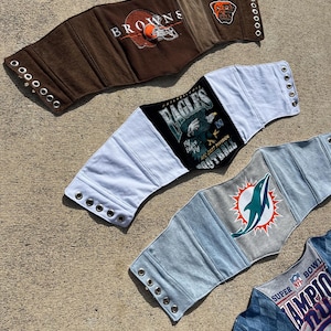Puede incluir: Cuatro cinturones estilo corsé confeccionados con ropa deportiva reutilizada. El cinturón superior es marrón con un logotipo de los Browns, el segundo es blanco y negro con un gráfico de los Eagles, el tercero es gris claro con un logotipo de los Dolphins y el inferior es azul y gris con texto del Super Bowl.