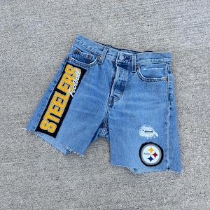 Könnte beinhalten: Hellblaue Jeansshorts mit Used-Details. Mit einem schwarzen und goldenen Aufnäher mit der Aufschrift "STEELERS Football" an der Seite. Enthält einen Steelers-Logo-Aufnäher und einen abgenutzten Bereich auf der Vorderseite.