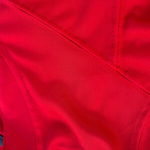 Peut inclure: Gros plan sur un maillot de sport rouge vif. Le maillot pr&eacute;sente des d&eacute;tails brod&eacute;s blancs et jaunes, avec des coutures et des coutures visibles. Le tissu semble &ecirc;tre un mat&eacute;riau synth&eacute;tique lisse.