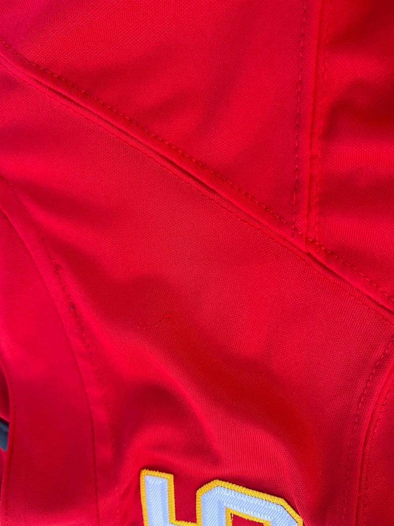 Peut inclure: Gros plan sur un maillot de sport rouge vif. Le maillot pr&eacute;sente des d&eacute;tails brod&eacute;s blancs et jaunes, avec des coutures et des coutures visibles. Le tissu semble &ecirc;tre un mat&eacute;riau synth&eacute;tique lisse.