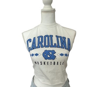 Op de afbeelding: Witte crop top met het woord "CAROLINA" in blauwe blokletters. Daaronder een blauw logo en het woord "BASKETBALL" in zwart. De top wordt op een mannequin gepresenteerd.