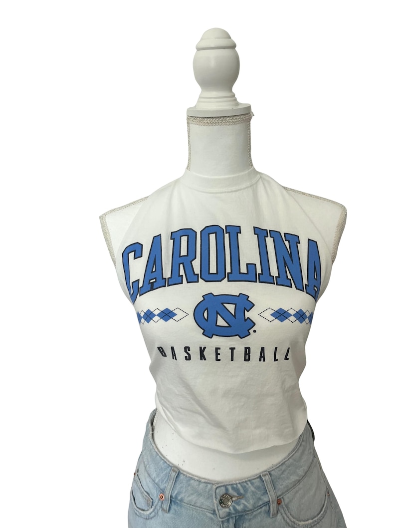 Op de afbeelding: Witte crop top met het woord "CAROLINA" in blauwe blokletters. Daaronder een blauw logo en het woord "BASKETBALL" in zwart. De top wordt op een mannequin gepresenteerd.