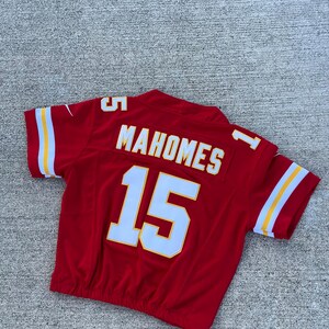 Peut inclure: Un maillot de football rouge avec des d&eacute;tails blancs et jaunes. Le maillot porte le nom "MAHOMES" et le num&eacute;ro "15" en lettres blanches. Les manches ont des rayures blanches avec une bordure jaune.