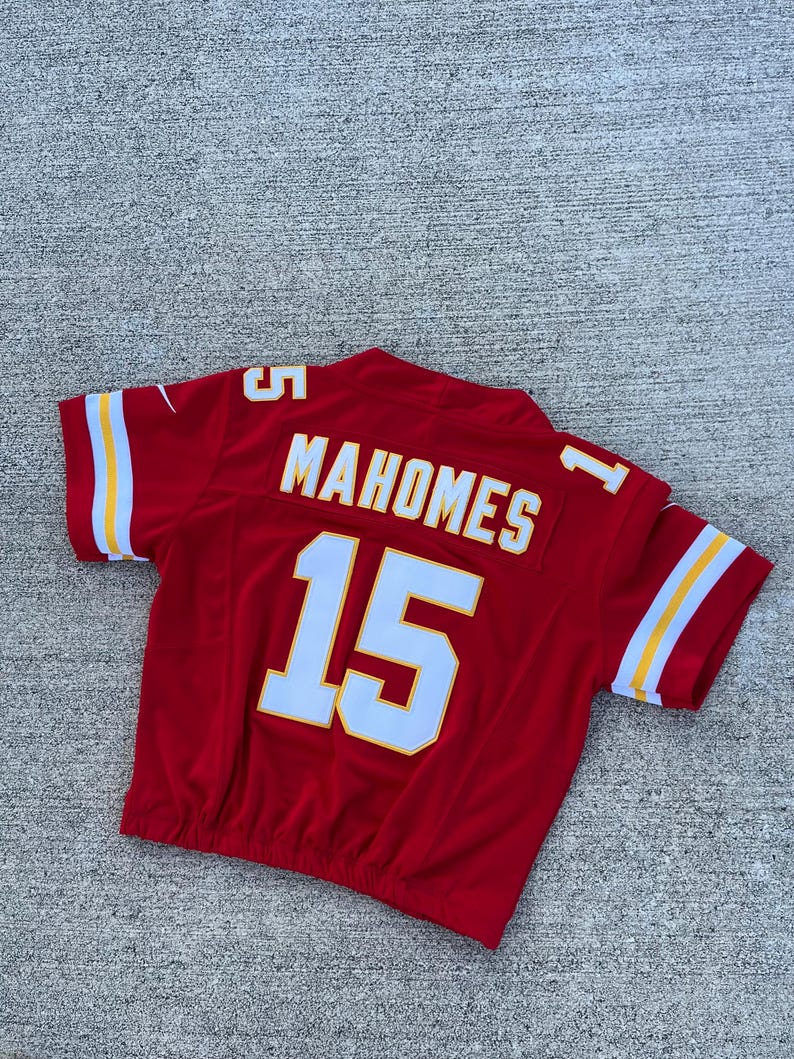 Peut inclure: Un maillot de football rouge avec des d&eacute;tails blancs et jaunes. Le maillot porte le nom "MAHOMES" et le num&eacute;ro "15" en lettres blanches. Les manches ont des rayures blanches avec une bordure jaune.