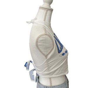 Op de afbeelding: Witte crop top met blauwe tekst en ruitpatroon. De top heeft striksluitingen op de schouders en taille. De top wordt gecombineerd met een lichtblauwe jeans.