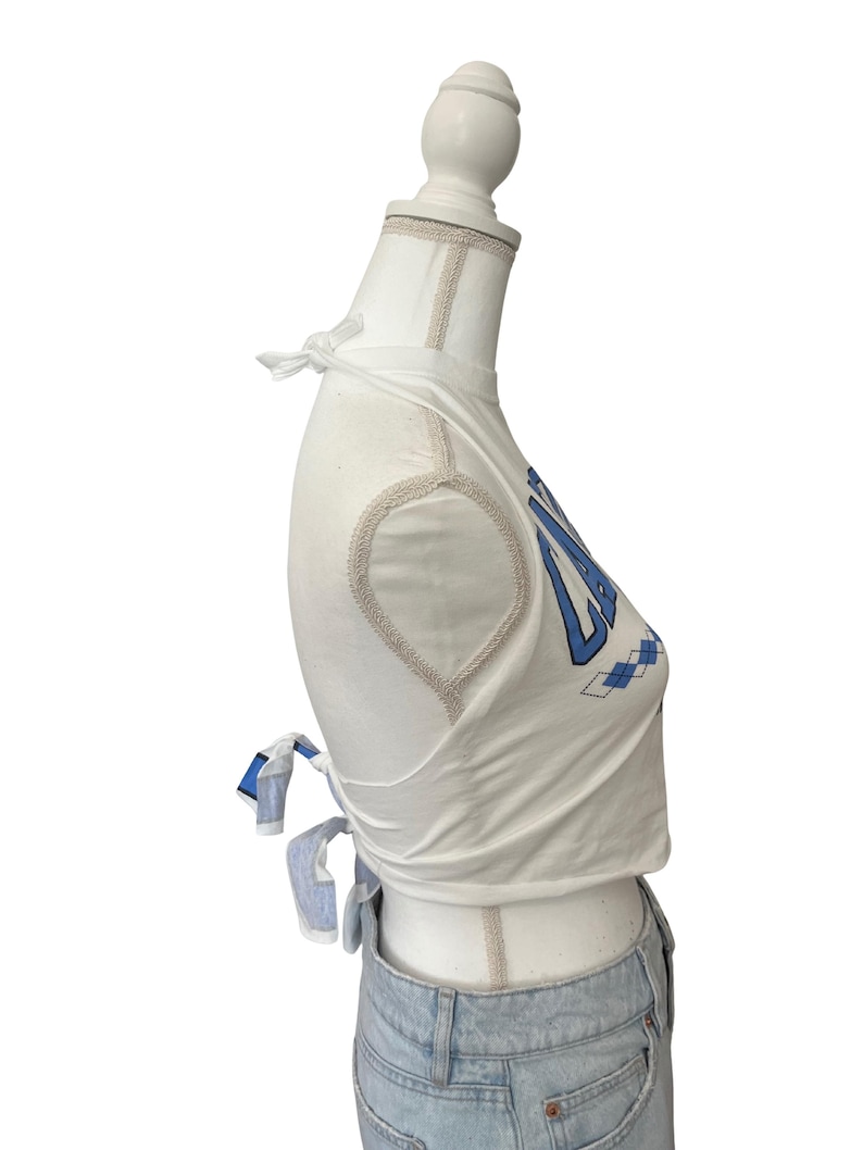 Op de afbeelding: Witte crop top met blauwe tekst en ruitpatroon. De top heeft striksluitingen op de schouders en taille. De top wordt gecombineerd met een lichtblauwe jeans.