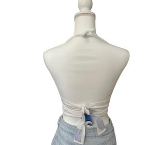 Op de afbeelding: Een witte haltertop met een striksluiting aan de achterkant. De top wordt getoond op een mannequin en gecombineerd met een lichtblauwe spijkerbroek. De stropdas heeft een blauw-wit patroon.