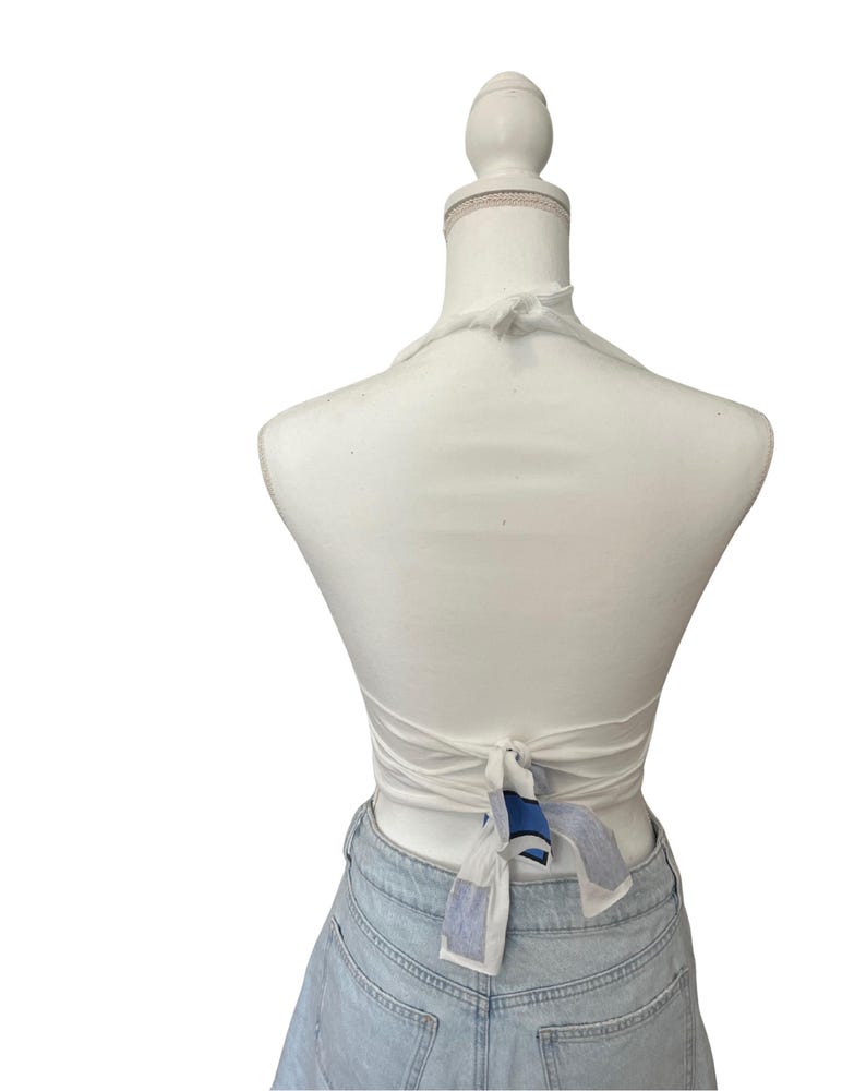 Op de afbeelding: Een witte haltertop met een striksluiting aan de achterkant. De top wordt getoond op een mannequin en gecombineerd met een lichtblauwe spijkerbroek. De stropdas heeft een blauw-wit patroon.
