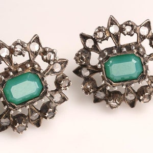 May include: A pair of silver earrings with a green stone set in a floral design. The earrings are surrounded by smaller clear stones.