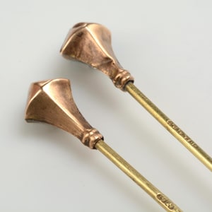 May include: Two gold-toned metal hat pins with a square-shaped top. The pins are about 10 inches long.