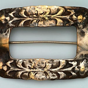 May include: A rectangular, antique silver-toned buckle with floral and leaf designs. The buckle has a hammered texture and a pin closure on the back. The buckle's design features a central opening and is likely for a belt or other garment.