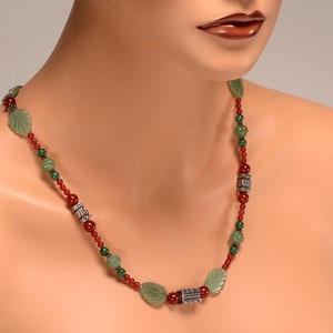 May include: A necklace featuring green leaf-shaped beads, red beads, and silver-colored cylindrical beads. The necklace is displayed on a mannequin. The beads are strung together with a red string.