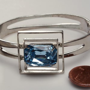 May include: A silver bangle bracelet with a square frame holding a large, light blue, faceted gemstone. The bracelet is open and the gemstone is centered in the frame.