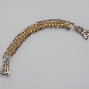 May include: A vintage bracelet featuring a curved design with three rows of small, round, cream-colored beads. The beads are connected by a delicate chain. The bracelet has ornate, silver-toned clasps on each end.