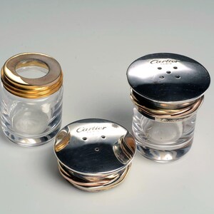May include: Three glass salt and pepper shakers with silver lids and gold bands. The lids have the word 'Cartier' engraved on them.