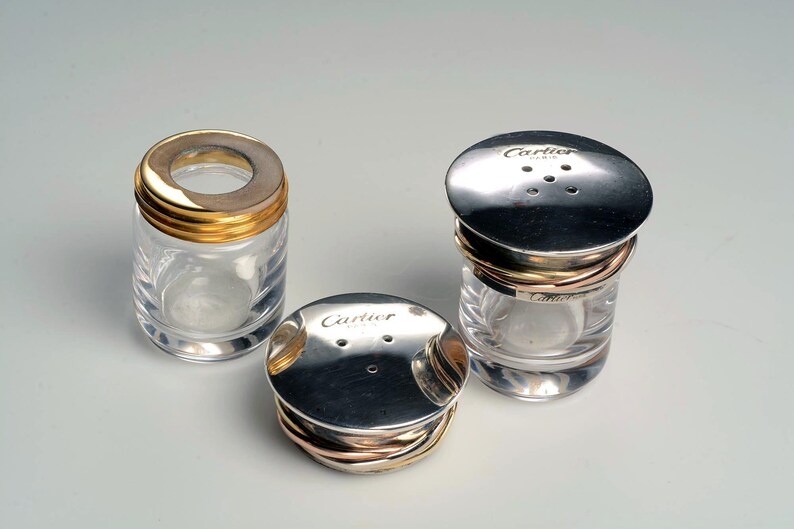 May include: Three glass salt and pepper shakers with silver lids and gold bands. The lids have the word 'Cartier' engraved on them.