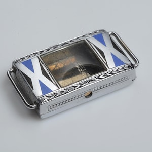 May include: A rectangular, silver-toned metal cigarette case with a hinged lid. The lid features blue and white cross designs. The case has a clear window and a small, circular opening on the side. The case has a decorative border.