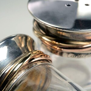 May include: Two silver salt and pepper shakers with gold and copper bands. The shakers are made of glass and have a clear glass body. The shakers are labeled 'Cartier France'.