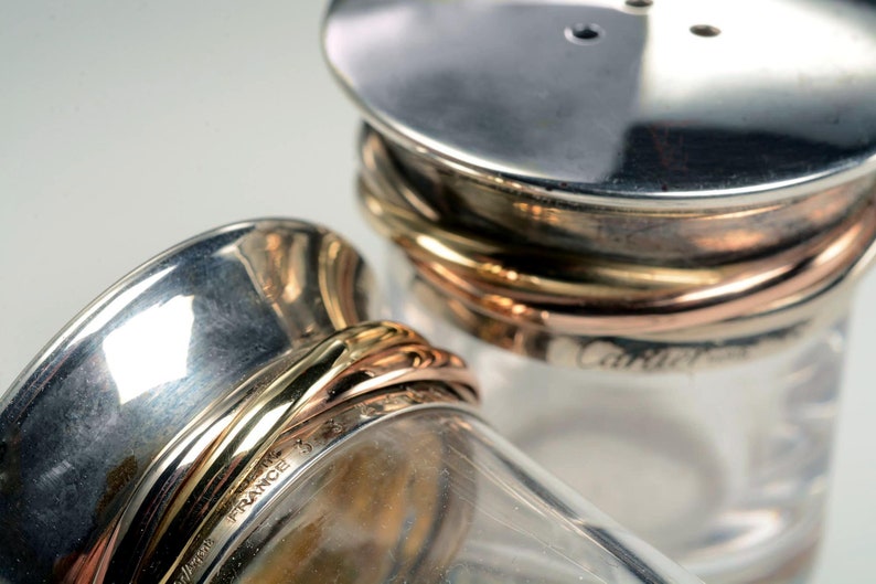 May include: Two silver salt and pepper shakers with gold and copper bands. The shakers are made of glass and have a clear glass body. The shakers are labeled 'Cartier France'.