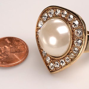 May include: A gold-toned ring with a large, pearly white center stone, surrounded by a border of clear, sparkling crystals. The ring is set on a wide band. A penny is next to the ring for size comparison.