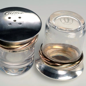 May include: Two clear glass salt and pepper shakers with silver lids and gold bands. The lid of the salt shaker has the words 'Cartier Paris' engraved on it. The lid of the pepper shaker has a stylized 'C' engraved on it.