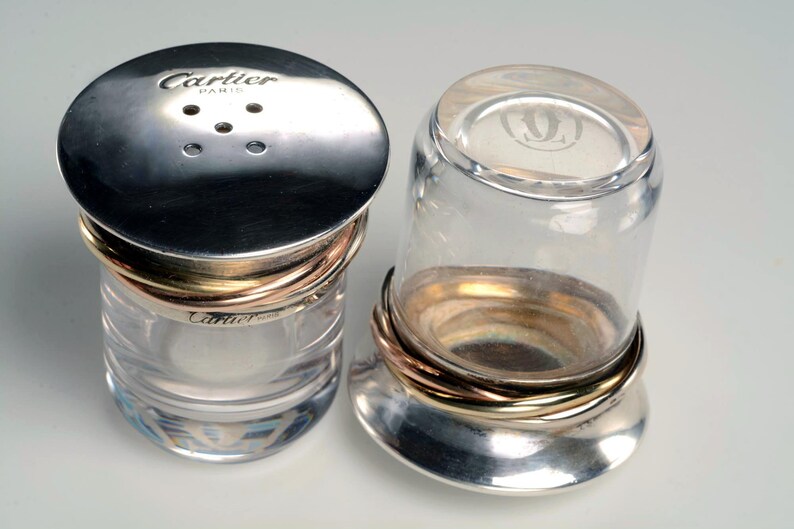 May include: Two clear glass salt and pepper shakers with silver lids and gold bands. The lid of the salt shaker has the words 'Cartier Paris' engraved on it. The lid of the pepper shaker has a stylized 'C' engraved on it.