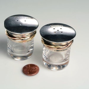 May include: Two clear glass salt and pepper shakers with silver lids and gold bands. The lids have the word 'Cartier' engraved on them.