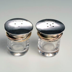 May include: Two silver salt and pepper shakers with gold and rose gold bands. The shakers have a clear glass base and a silver lid with the word 'Cartier' engraved on the top.