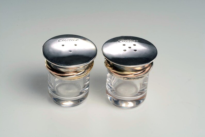 May include: Two silver salt and pepper shakers with gold and rose gold bands. The shakers have a clear glass base and a silver lid with the word 'Cartier' engraved on the top.