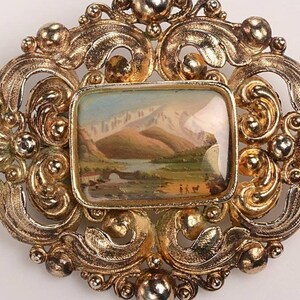 May include: Vintage gold-tone brooch with an oval filigree design. The center features a rectangular miniature landscape painting of mountains, a lake, and figures. A classic accessory.