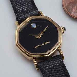 May include: A gold-toned, octagonal wristwatch with a black face and a black leather band. The watch face has gold hands and the text "NEIMAN-MARCUS". A small, iridescent stone is set at the 12 o'clock position. A penny is in the background.