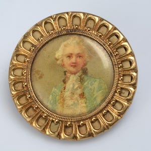 May include: A gold-toned round brooch with a raised, ornate frame. The center features a portrait of a man with a white powdered wig, wearing a blue and white patterned jacket.