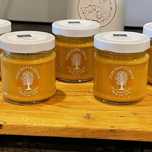 May include: Five jars of Pure Maple Cream with white lids and labels featuring a tree logo. The jars are filled with a golden-colored cream and are displayed on a wooden cutting board. Each jar contains 4 fl oz.