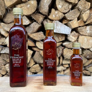May include: Three glass bottles of pure Vermont maple syrup in different sizes. The bottles are labeled with the text "Pure Vermont Maple Syrup" and the size in milliliters and fluid ounces. The bottles are sitting on a wooden surface in front of a stack of firewood.