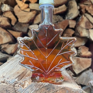 May include: A clear glass bottle shaped like a maple leaf, filled with amber liquid. The bottle has a white and gold cap. The bottle is sitting on a piece of wood, with a background of stacked firewood.