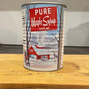 May include: A can of Pure Maple Syrup, 540 ml, with a red label. The label features a winter scene with a red barn, trees, and snow. The can is sitting on a wooden surface. The text "REFRIGERATE AFTER OPENING" is also visible.